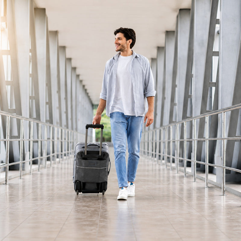 A man walking on a modern covered walkway while pulling a dark gray rolling suitcase. He is wearing a light blue button-up shirt over a white t-shirt, light blue jeans, and white sneakers.