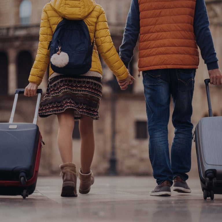 A couple walking away from the camera, holding hands and each pulling a wheeled suitcase. The woman wears a yellow jacket, a patterned skirt, and a navy backpack, while the man wears an orange puffer vest over a dark shirt and jeans.