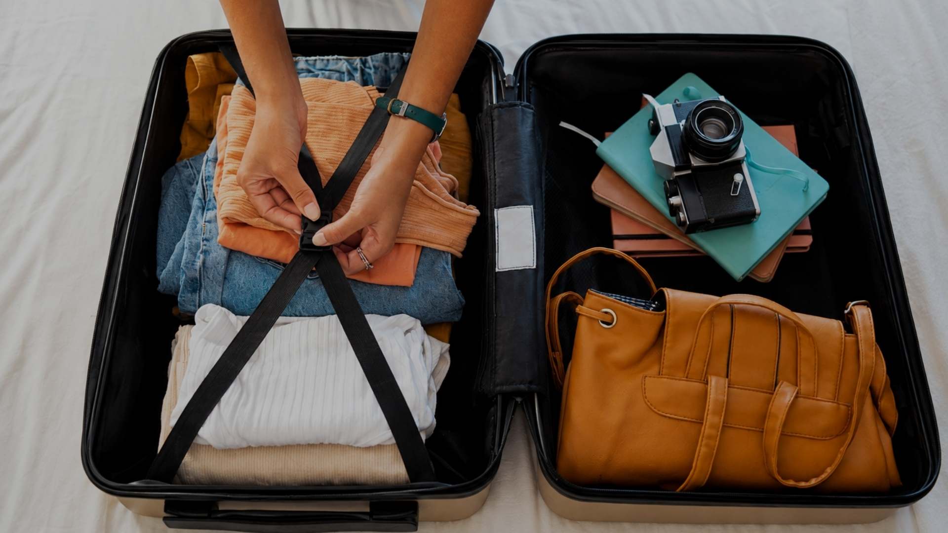An open black suitcase on a white bed, filled with folded clothes, a tan leather handbag, a teal notebook, a camera, and other travel items. A person's hands are visible adjusting the suitcase strap.
